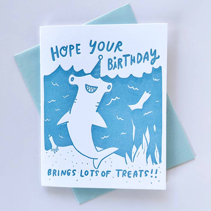 Greeting card with white background and blue ocean with a white shark wearing a blue party hat.  Blue text says, "Hope your birthday brings lots of treats!!". Blue envelope included. 