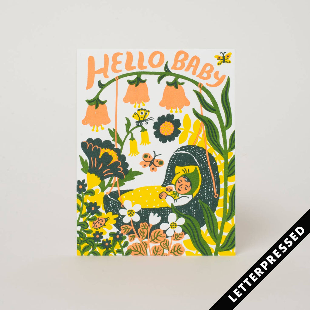 White background with image of baby in a green basket bassinet surrounded with yellow and orange flowers. Peach text says, "Hello Baby". Kraft envelope included.