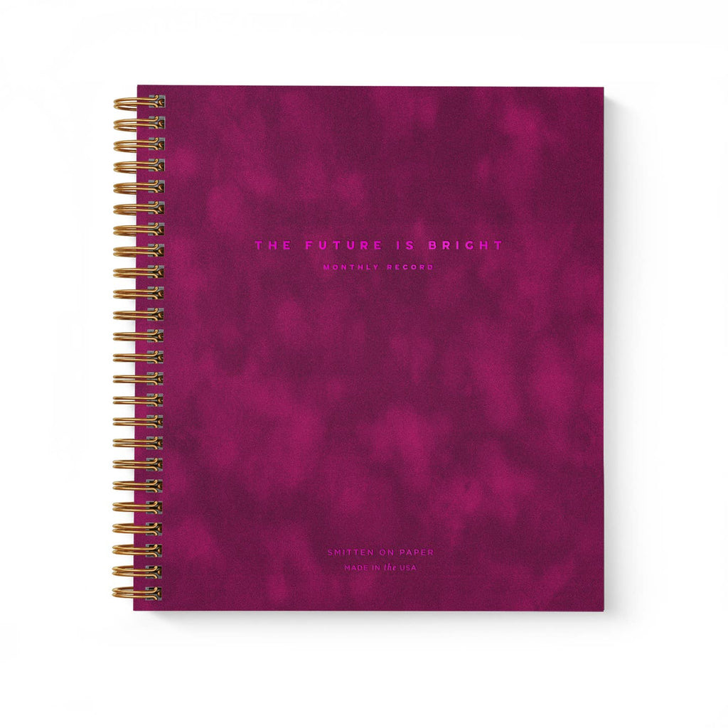 Planner with burgundy cover and hot pink text says, "The Future is bright, monthly planner" with gold coil binding on side.