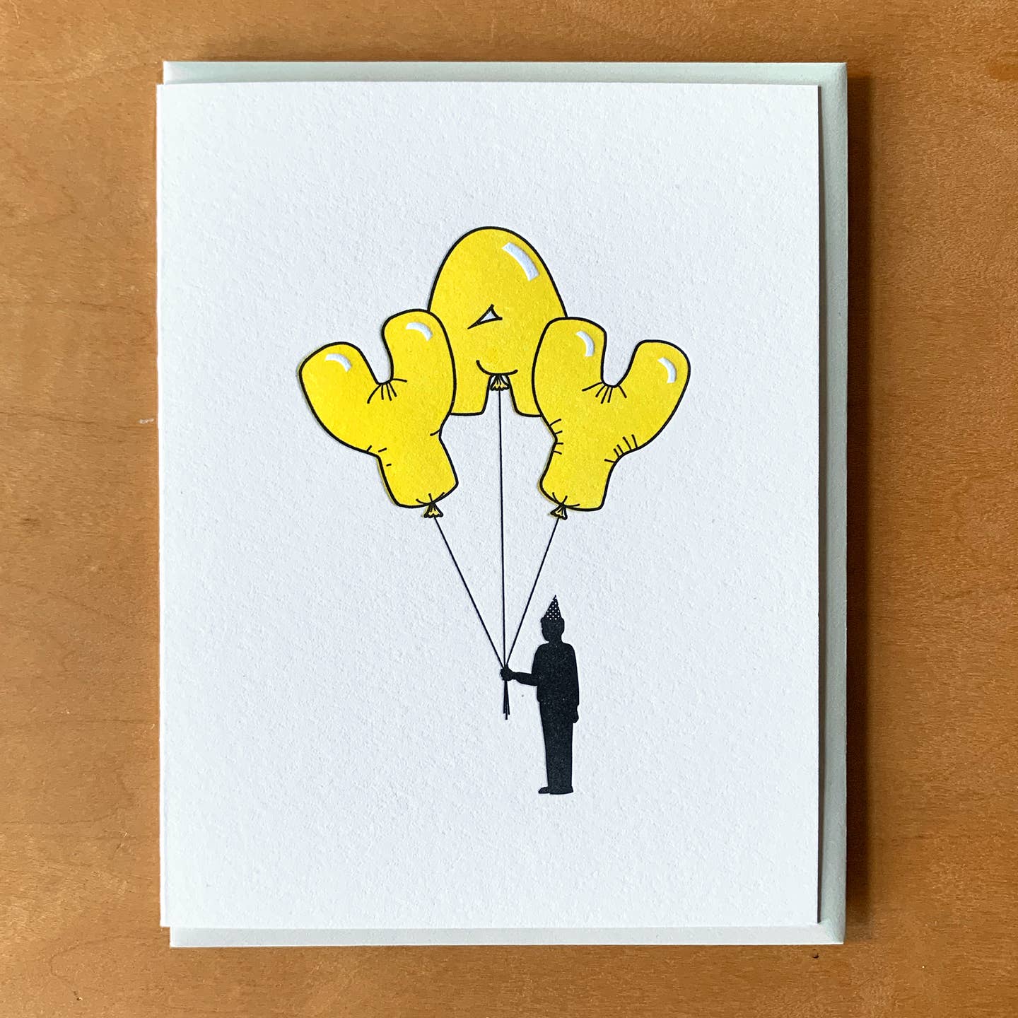 Greeting card with white background and image of yellow balloons "YAY" held by a black silhoutte of a person wearing a party hat. Grey envelope included. 