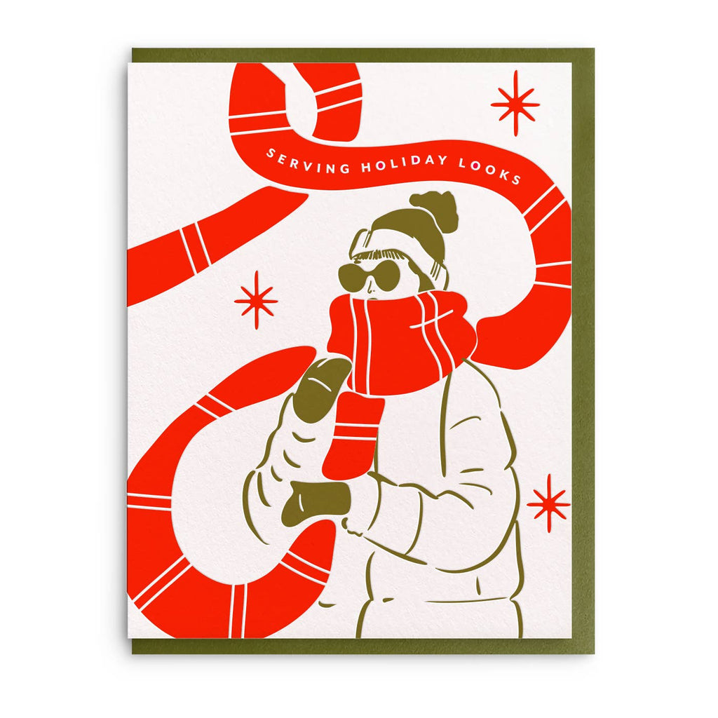 Greeting card with white background and iamge of a person in a white winter coat with green mittens and hat and a very long red scarf. White text on scarf says, "Serving holiday looks". Envelope included.