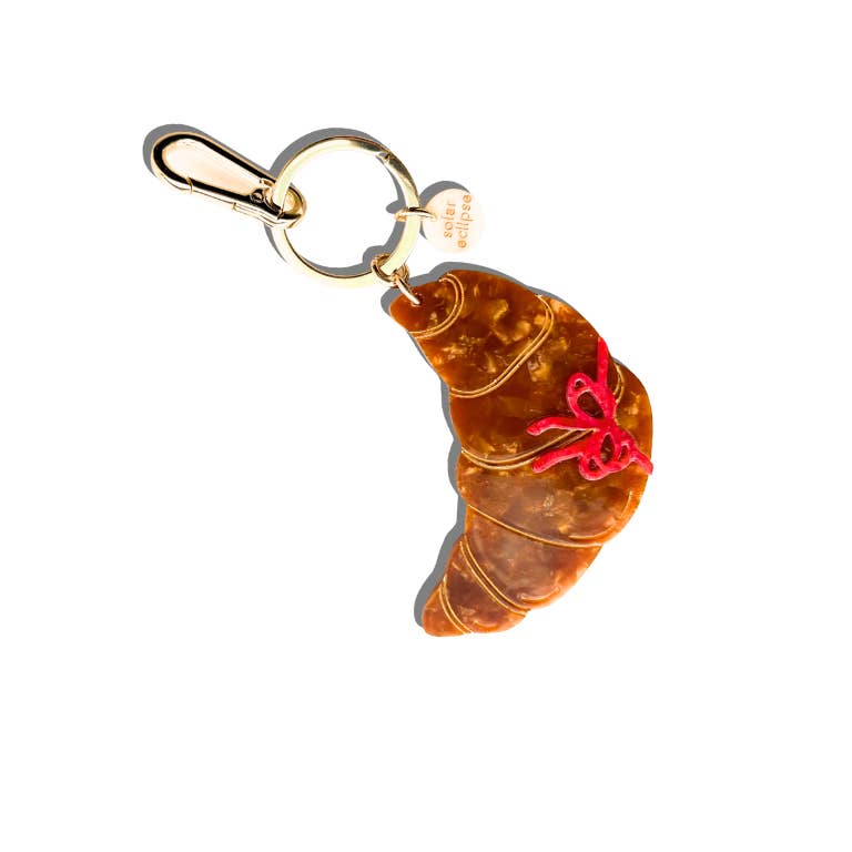 Image of keychain charm in the shape of a golden croissant with a red bow Gold colored key ring and clip. 