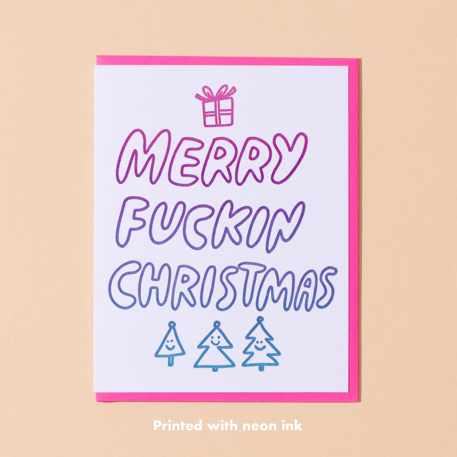 Image of card with white background and gradient text in pink to purple says, "Merry Fuckin Christmas" with image of gift and trees. Hot pink envelope included. 