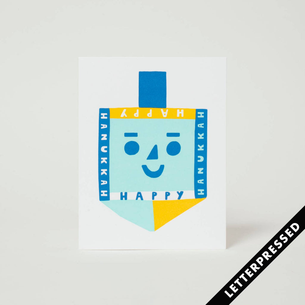 White background with image of blue and yellow dreidel  and white text says, "Happy Hanukkah". Kraft envelope included.