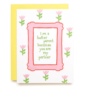 Greeting card with white background and images of small pink tulips and a frame of pink with green text says, "I am better parent because you are my partner". Yellow envelope included. 