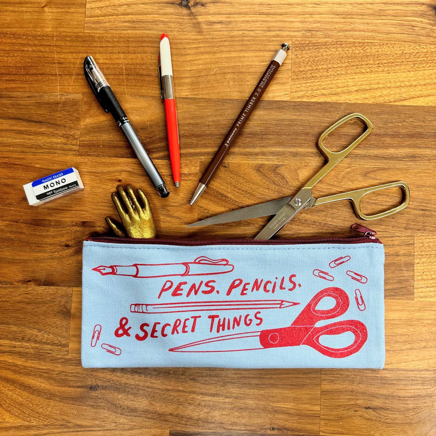Image of canvas zippered pencil case with cream background and images of fountain pen, pencil, scissors and paper clips in red. Red text says, "Pens, pencils & secret things". 