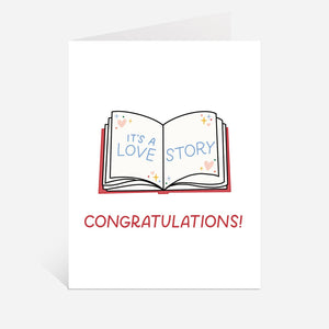 Greeting card with white background and image of an open book with blue text says, "It's a love story". Red text says, "Congratulations!". Envelope included.