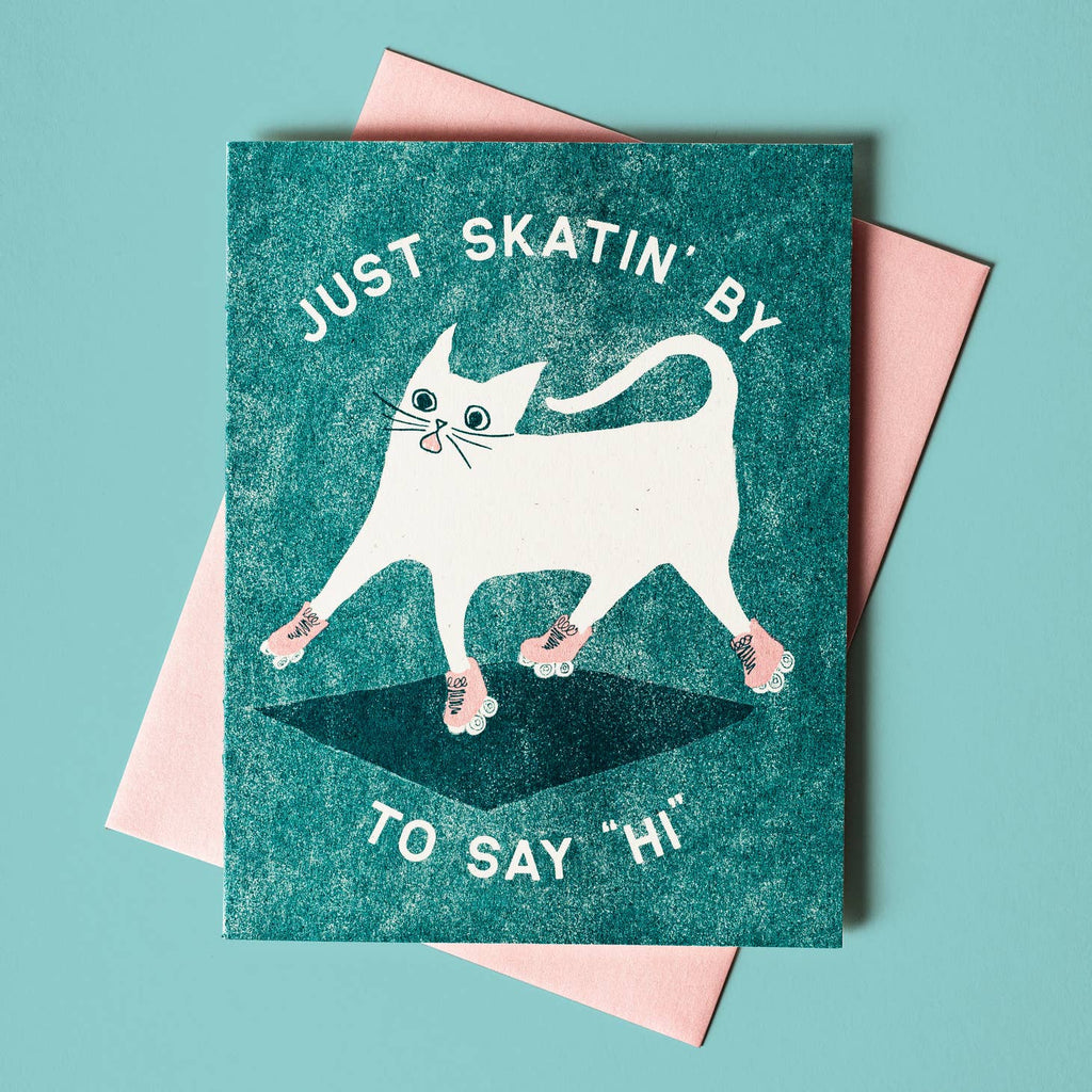 Teal background with image of white cat wearing pink roller skates and white text says, "Just skating' by to say Hi". Pink envelope is included. 