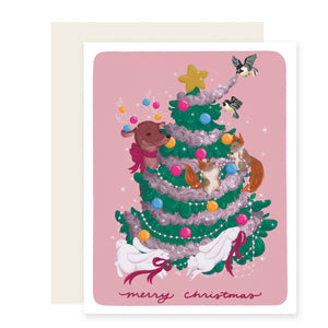 Greeting card with pink background and image of green tree with woodland creatures. Red text says, "Merry Christmas". Envelope included .