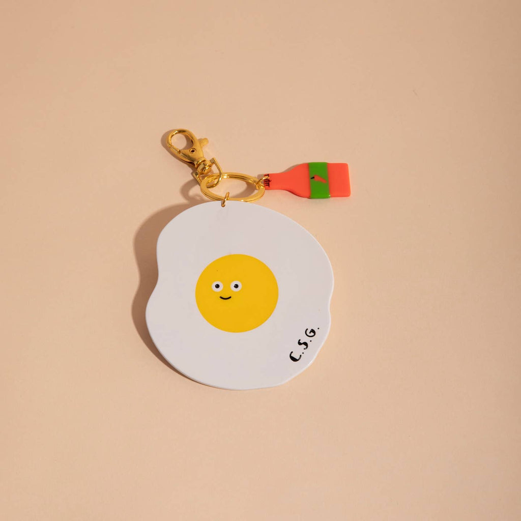 Image of a plastic charm in the shape of a fried egg and a bottle of hot sauce. 