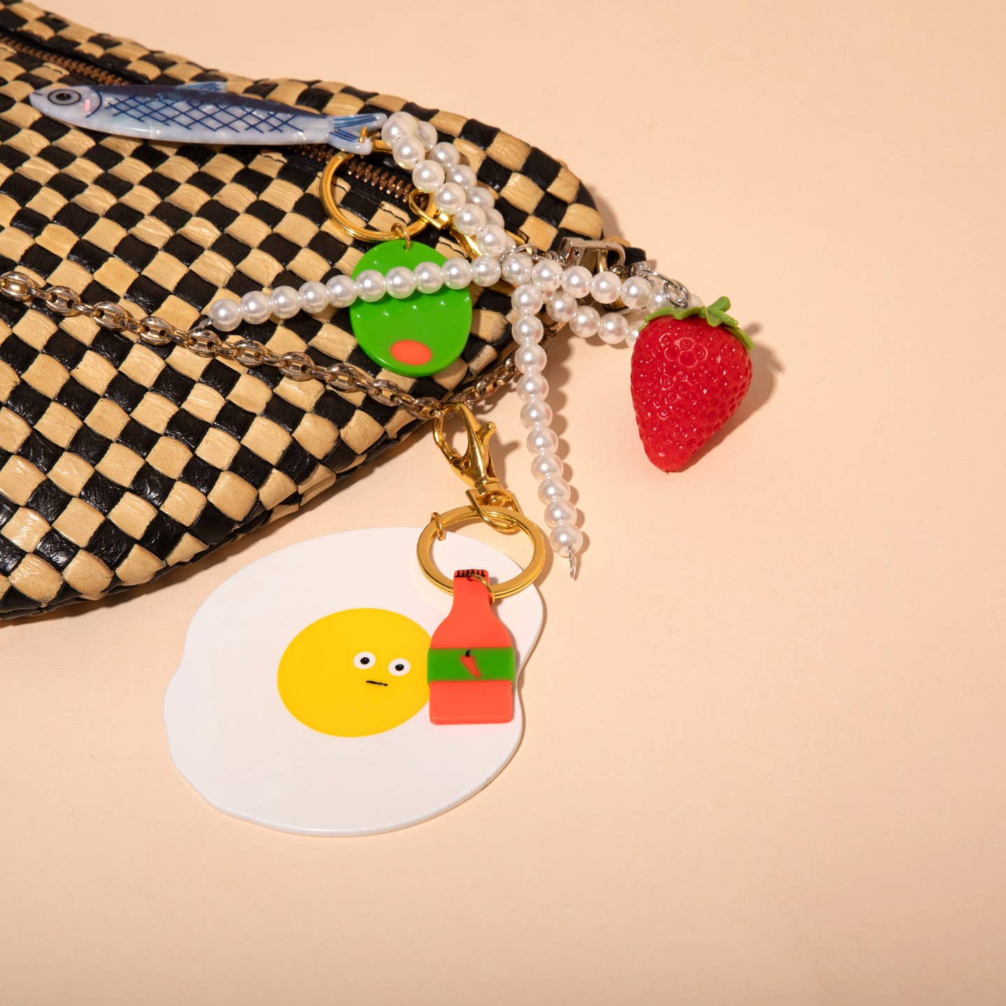 Image of a plastic charm in the shape of a fried egg and a bottle of hot sauce clipped to a checkered handbag.
