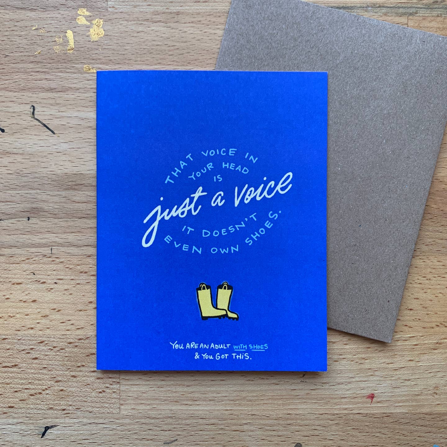 Greeting card with blue background with light blue and white text says, "That voice in your head is just a voice it doesn't even have shoes. You are an adult with shoes and you got this." with image of yellow rain boots. Grey envelope included.