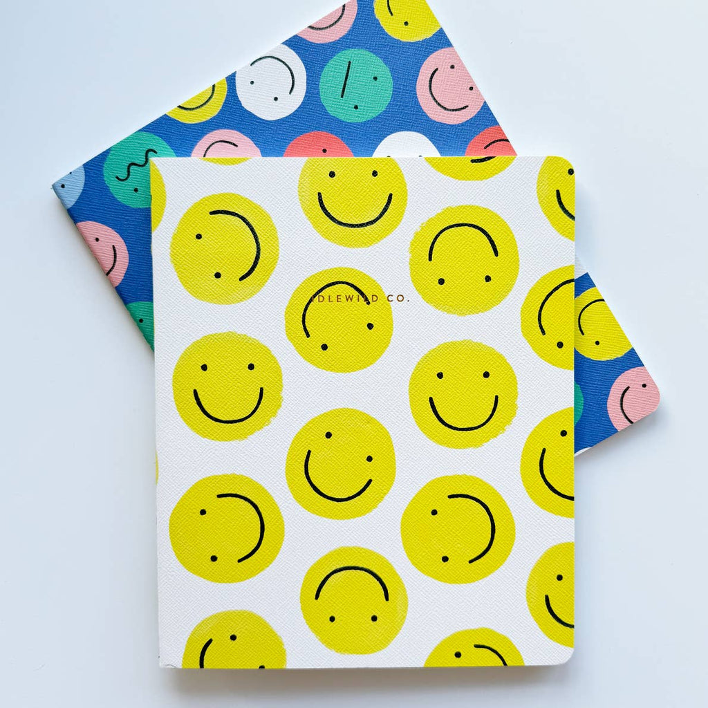 Image of two notebooks. One with white background with yellow smiley faces. One with blue background with images of pink, yellow, aqua, white and blue faces,