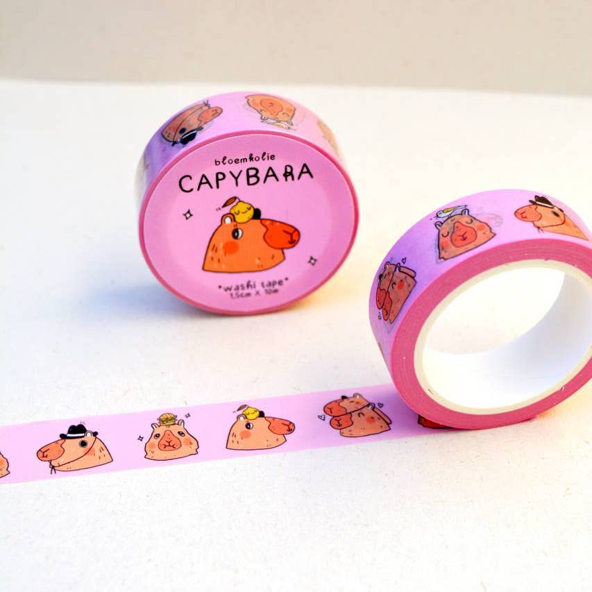 Image of roll of washi tape with pink background with images of capybaras in brown with orange notes dressed in hats and costumes.