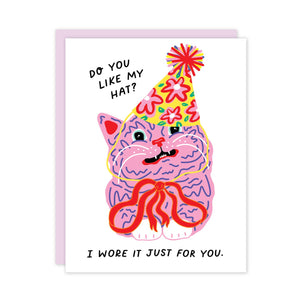 Greeting card with white background and an image of a pink cat with blue details and a red bow wearing a yellow flowered party hat. Black text say, "Do you like my hat? I wore it just for you.". Lilac envelope included.