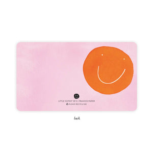 Back of little note is pale pink background with image of an orange smiley face on right side.