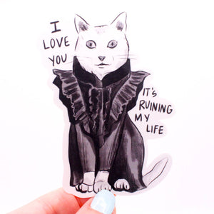 Image of sticker of a white cat wearing a black ruffles outfit with white background. Black text says, "I love you, it's ruining my life". 