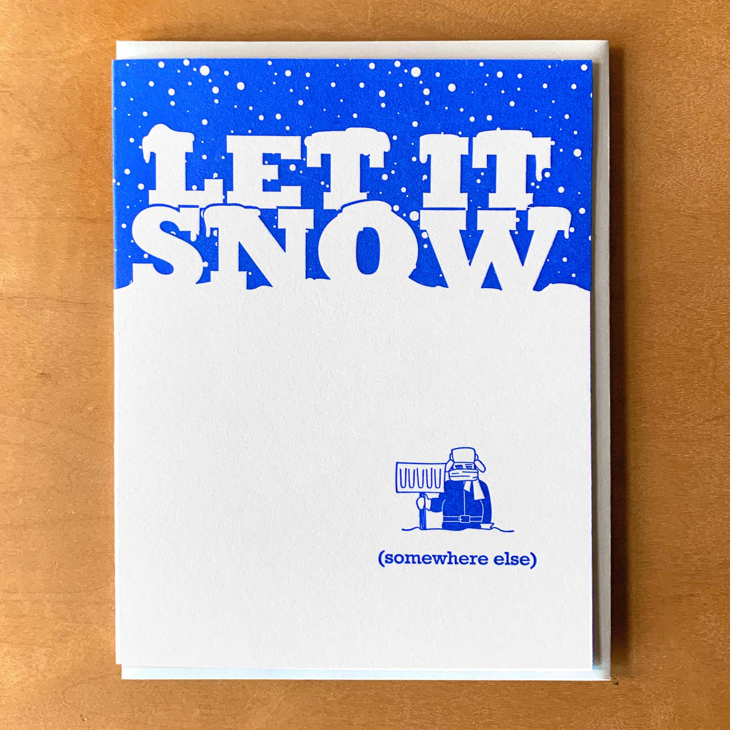 Greeting card with blue and white background and image of a person in layers of warm clothing holding a shovel. White text says, "Let it snow" and blue text says, "(Somewhere else)". Envelope included. 