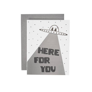 Greeting card with white background and image of a space ship with two aliens and a ray coming out from bottom. Black text says, "Here for you". Grey envelope included.