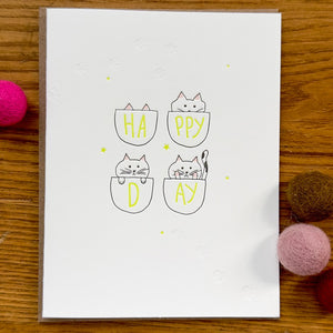 Greeting card with white background and images of four baskets with cats peering our of them. Yellow text says, "HA, PPY, D, AY" on the baskets. Envelope included. 