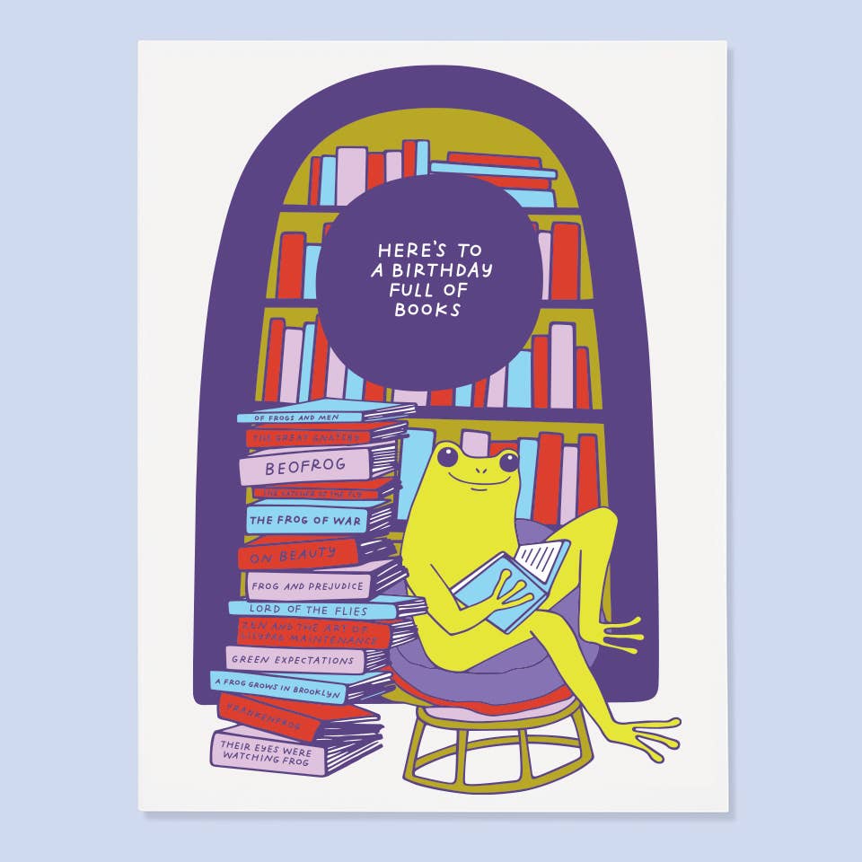 Greeting card with white background and image of a frog reading a book with a book shelf behind it and a stack of books. Purple text says, "Here's to a birthday full of books". Envelope included.