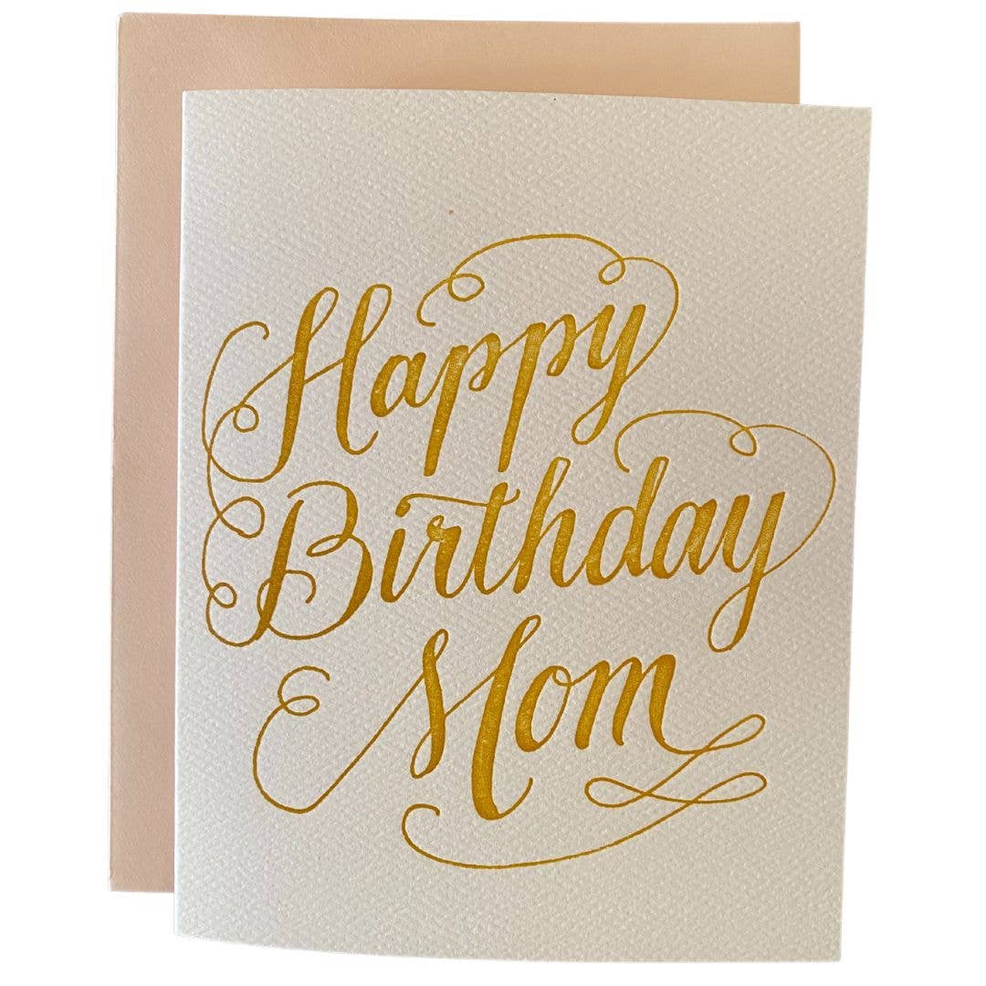 Greeting card with cream background with gold text says, "Happy Birthday Mom". Peach envelope included. 