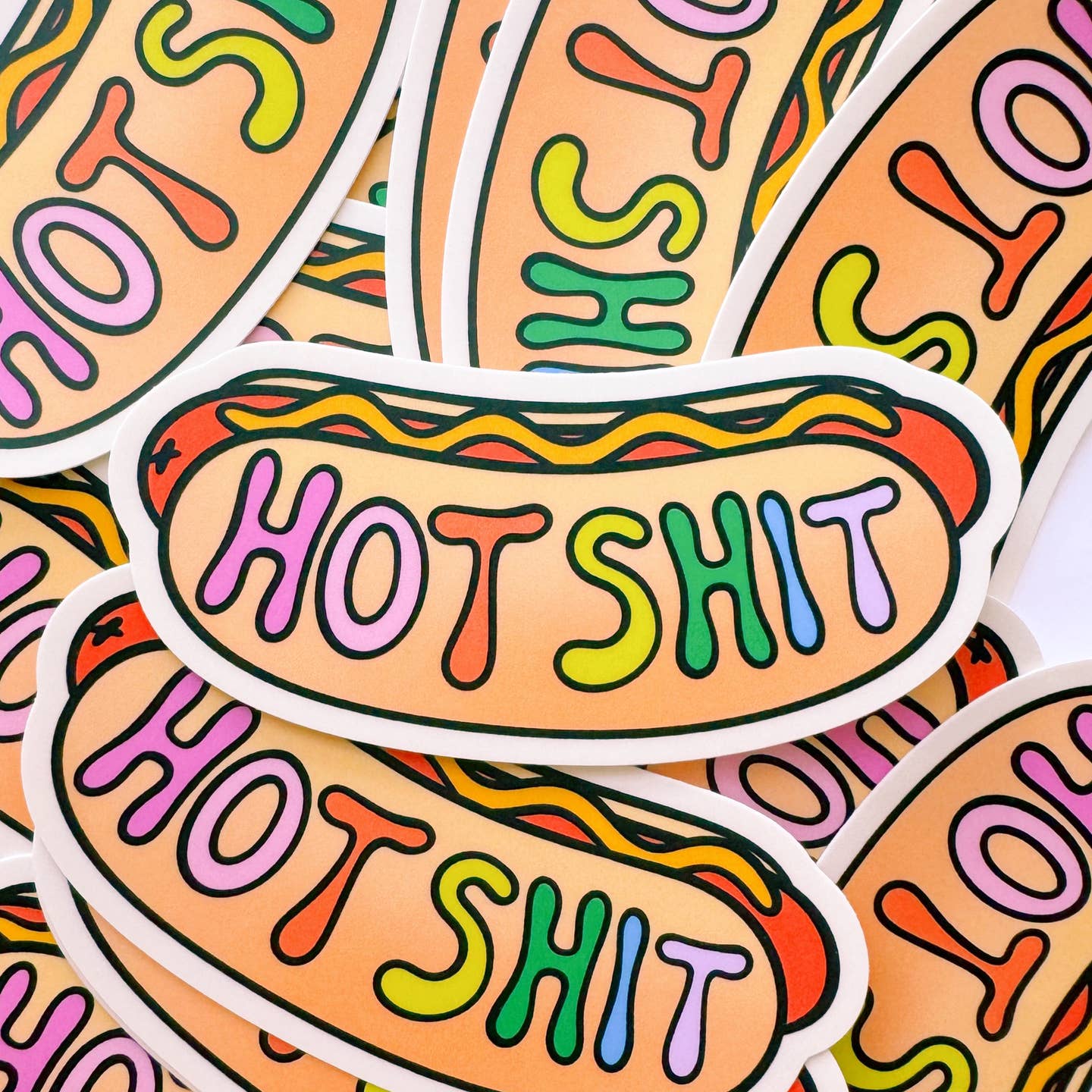 Decorative sticker in shape of a hot dog on a roll with drizzle of yellow mustard and rainbow text says, "Hot Shit". 