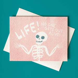 Greeting card with pink background and image of a white skeleton and white text says, "Life! No one gets out alive!". White envelope included.