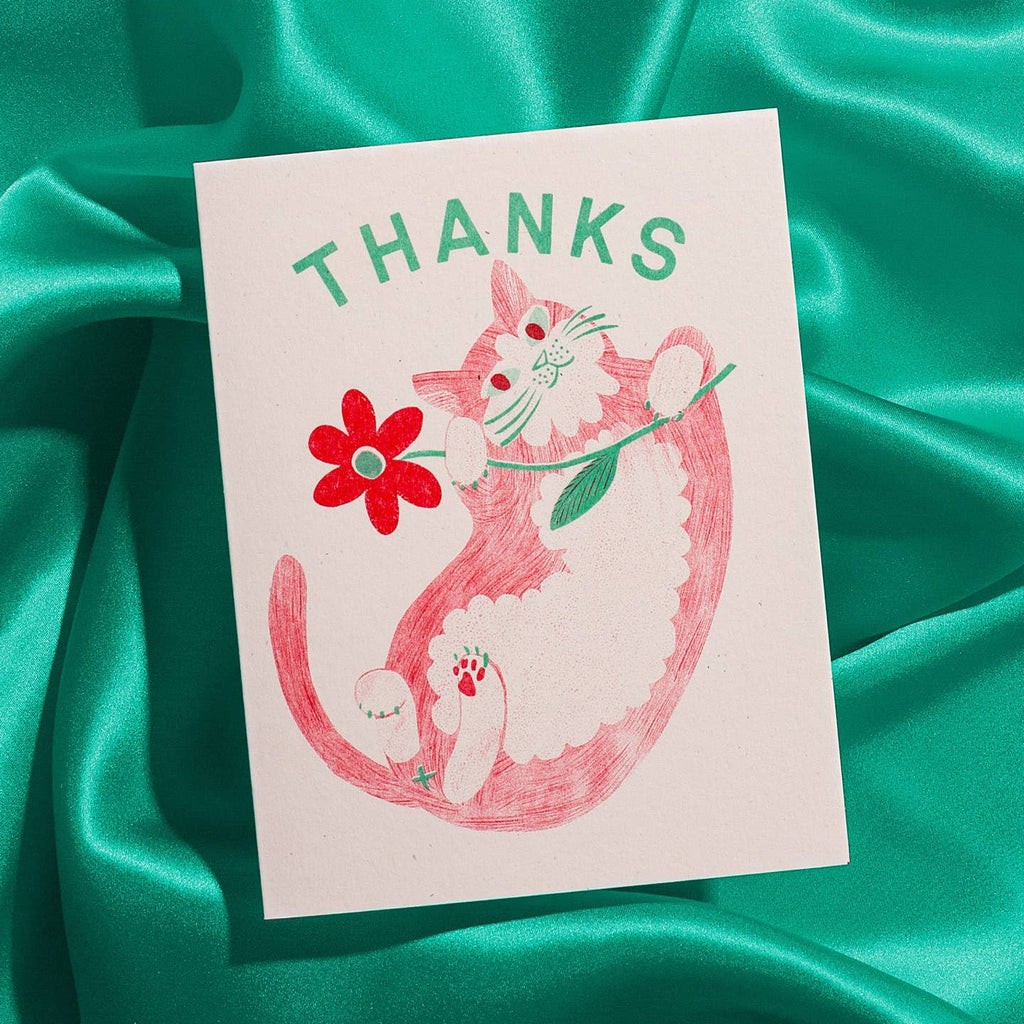 Greeting card with cream background and image of a red and white cat on its back holding a red flower and green text says, "Thanks". Envelope included.