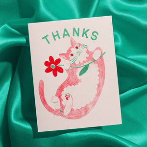 Greeting card with cream background and image of a red and white cat on its back holding a red flower and green text says, "Thanks". Envelope included.