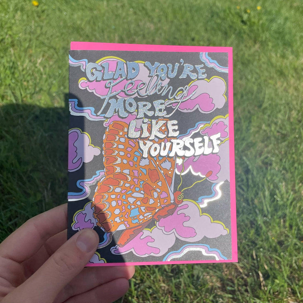 Image of card with black background with image of an orange and red butterfly with pink clouds and blue, pink, and white ribbons. Grey text says, “Glad you’re feeling more like yourself”. A bright pink envelope is included. 
