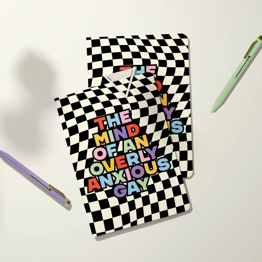 Image of notebook with black and white checkerboard patterned background with rainbow text says, "The mind of an overly anxious gay". 