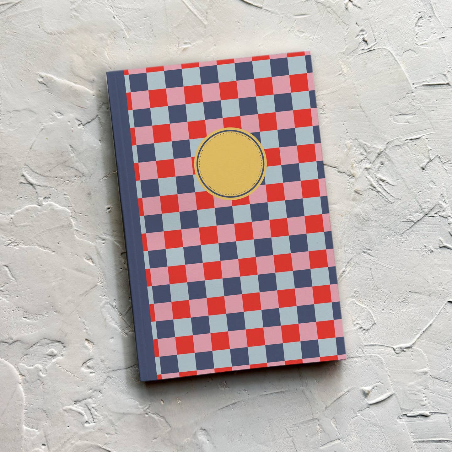 Notebook journal with blue, light blue, pink and red checkered background and navy binding with yellow circle in middle of front cover. 