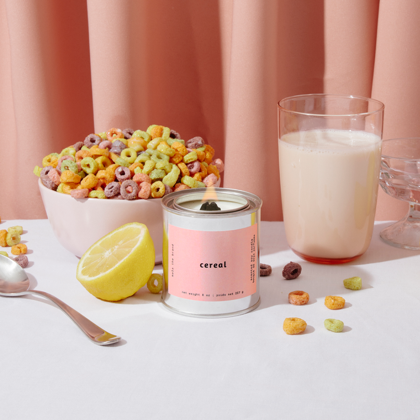 Image of candle with pink label and bowl of fruity cereal with glass of milk and lemon. 