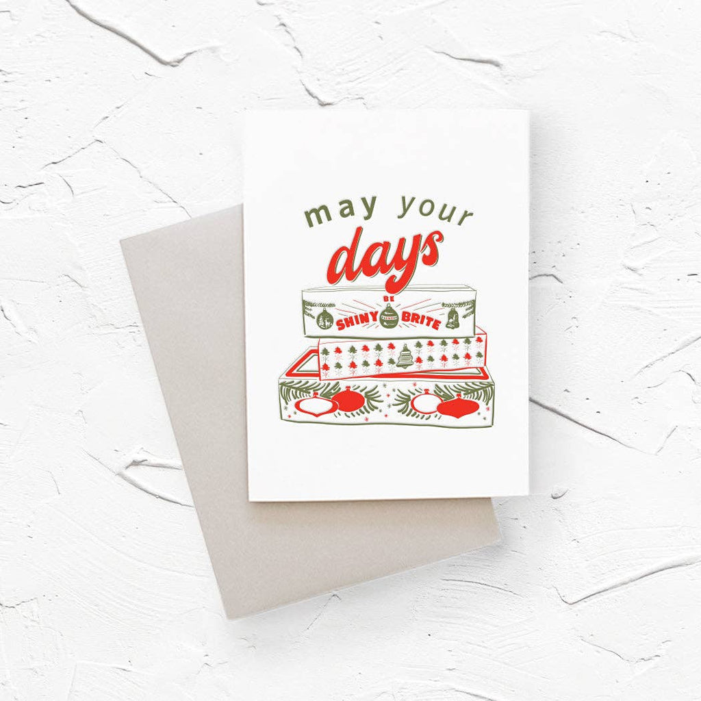 Greeting card with cream background and image of a stack of ornament boxes with green and red text says, "May you days be shiny and brite". Red envelope included. 