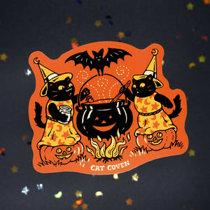 Decorative sticker with orange background and images of two black cats stirring a pumpkin shaped caldron. 