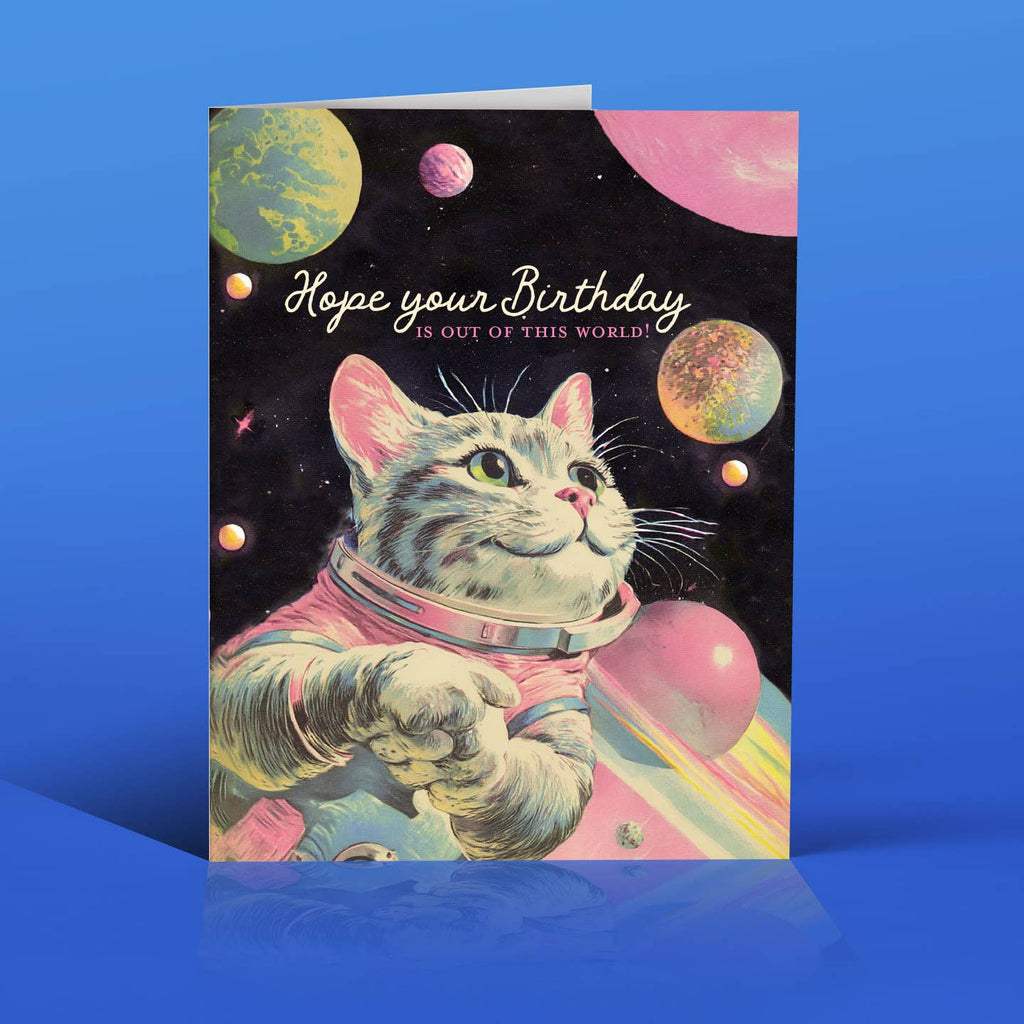 Greeting card with black background and planets and image of grey cat wearing space suit and white text says, "Hope your birthday" and pink text says, "Is out of this world!". Envelope included.