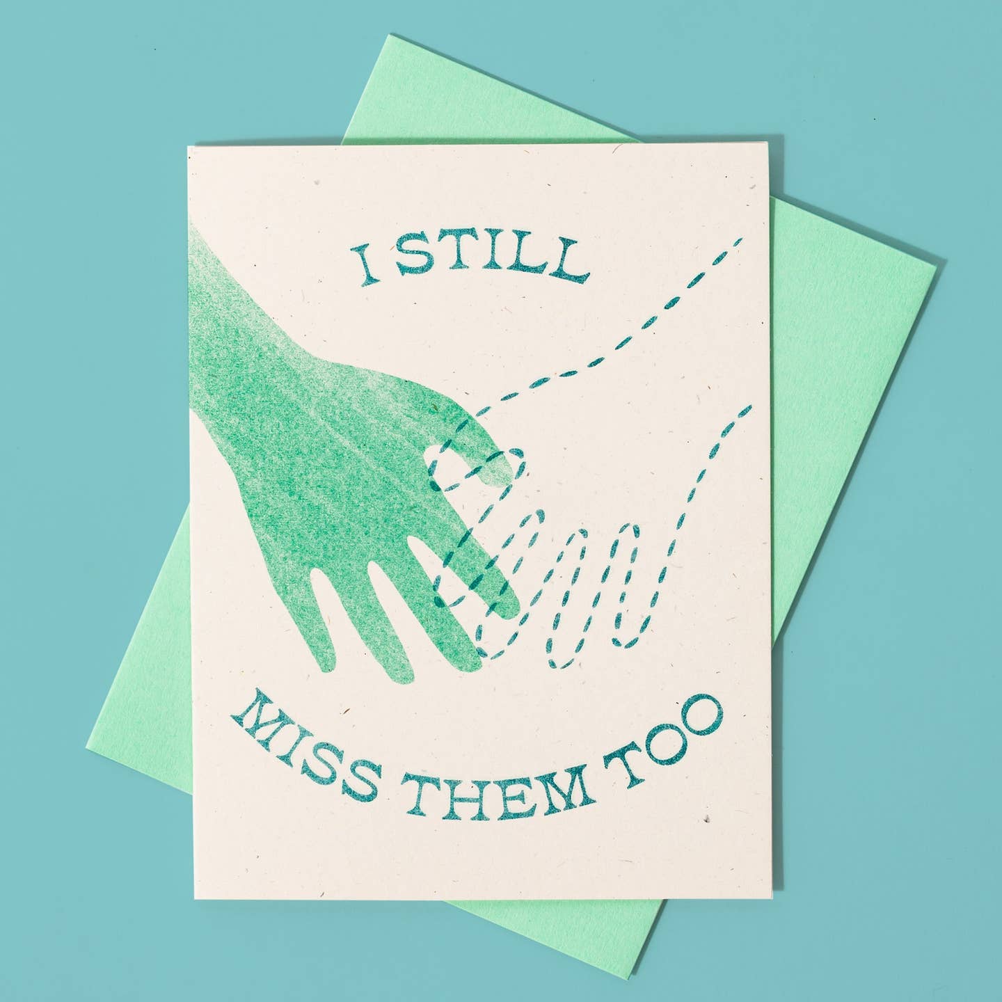 Greeting card with white background and image of two hands in aqua one in color and one with dotted line. Aqua text says, "I still miss them too". Aqua envelope included.