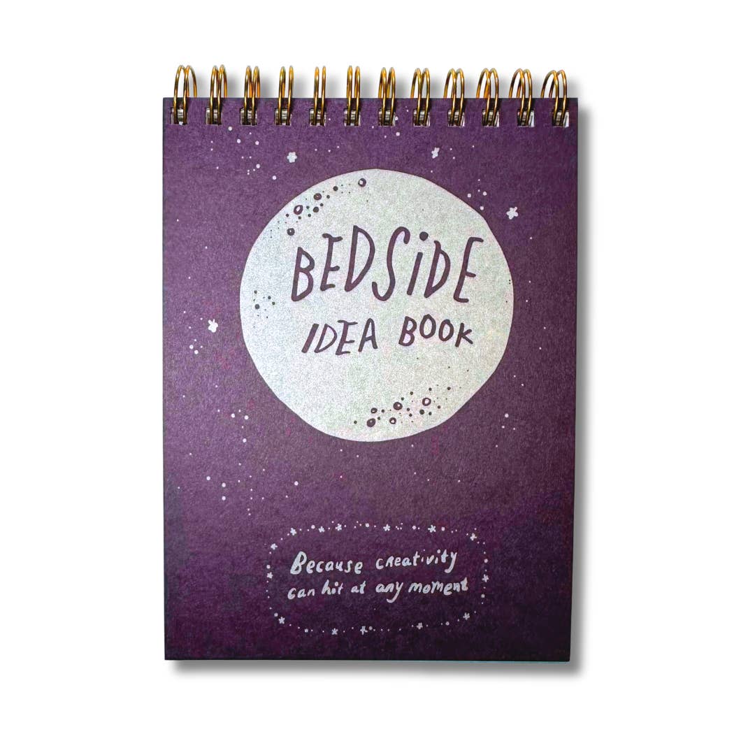Image of small notepad with purple background and silver circle with purple text says, "Bedside idea book" and silver text says, "Because creativity can hit at any moment". Coil binding on top.