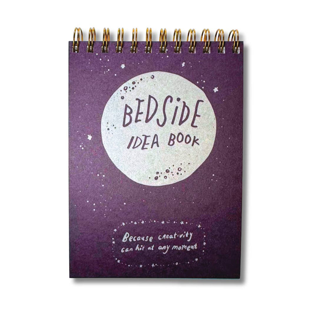 Image of small notepad with purple background and silver circle with purple text says, "Bedside idea book" and silver text says, "Because creativity can hit at any moment". Coil binding on top.