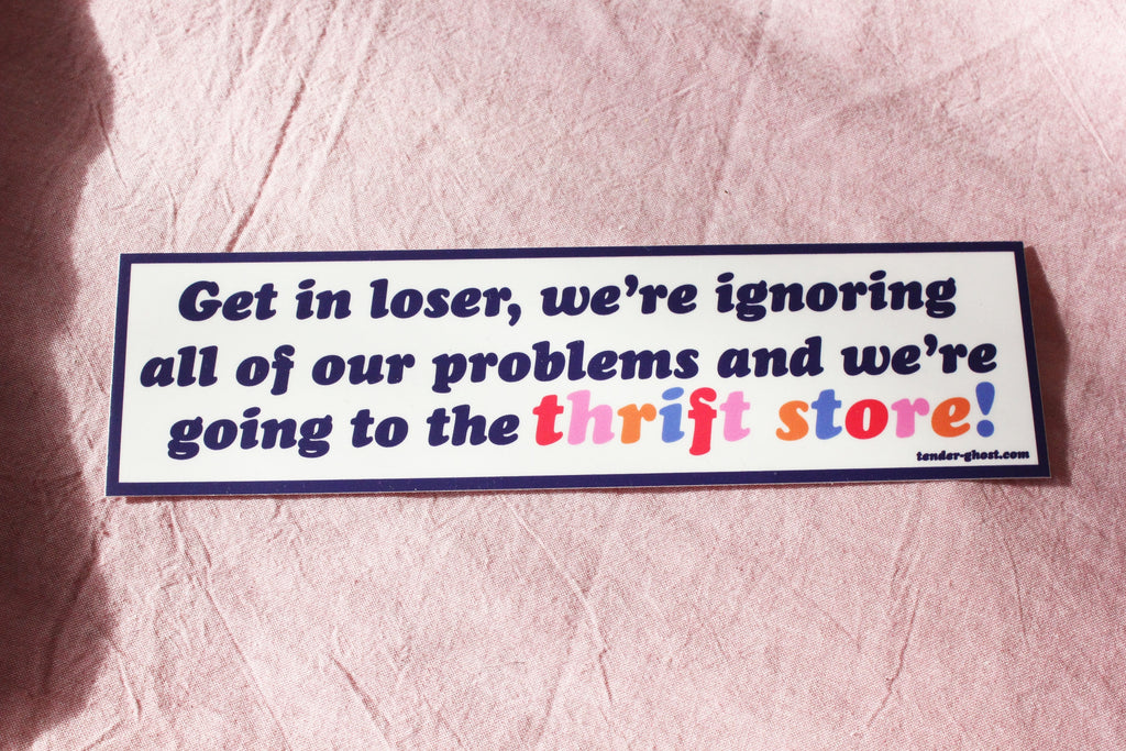 Bumper sticker with white background and black border with black text says, "Get in loser, we're ignoring all of our problems and we're going to the" and multicolored text says, "thrift store!". 