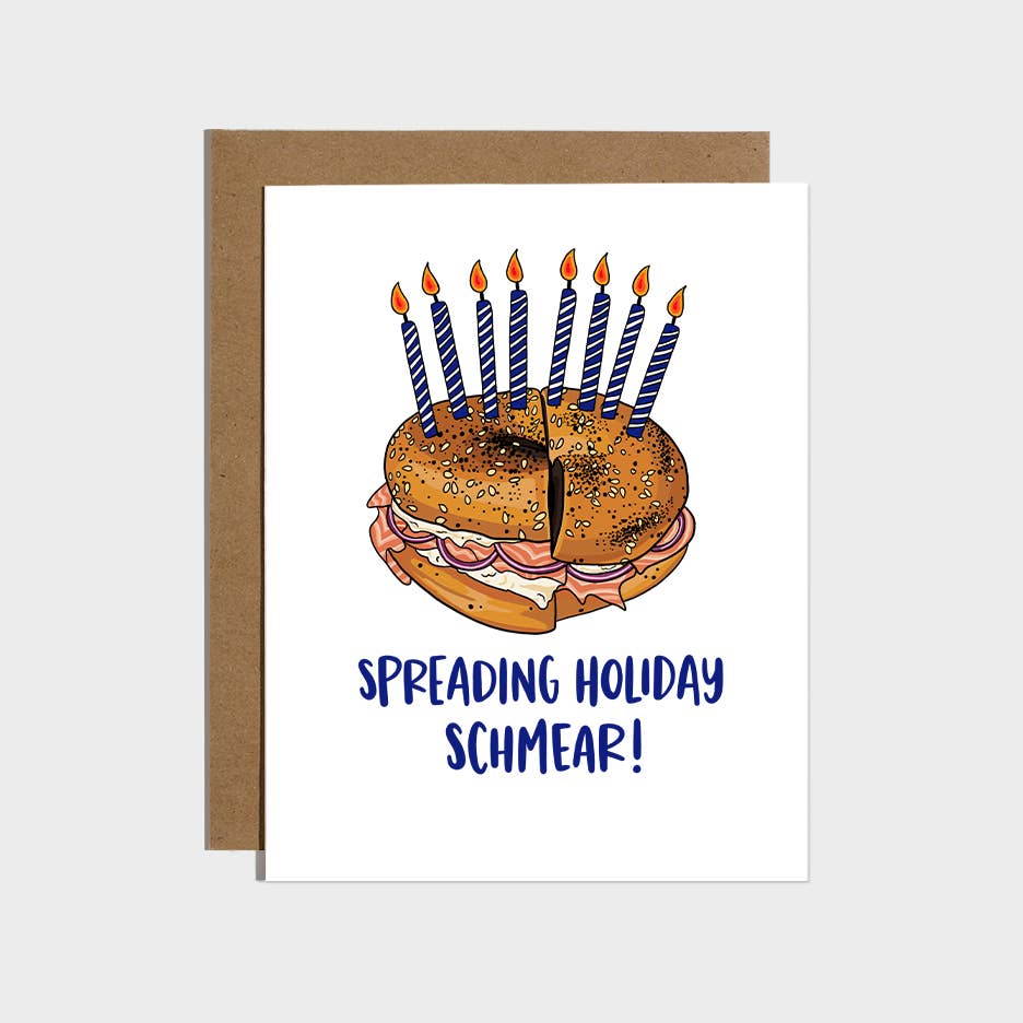 Greeting card with white background and image of  bagel with cream cheese and lox with Hanukkah candles and blue text says, "Spreading holiday schmear!". Kraft envelope included. 