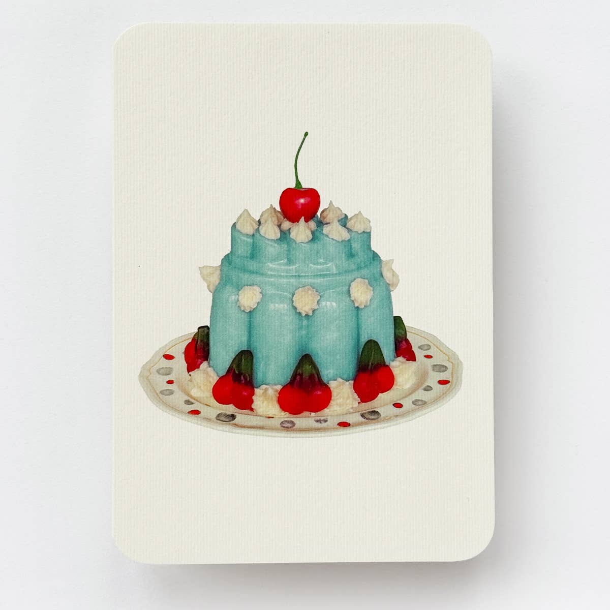 Greeting card with cream background and image of blue Blancmange dessert withj red cherries and white dollops of cream on a ivory plate. Envelope included.