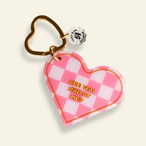 Image of keychain with heart shaped with pink and white checkerboard pattern and gold heart shaped ring for keys. Gold text says, "Are you mad at me?".