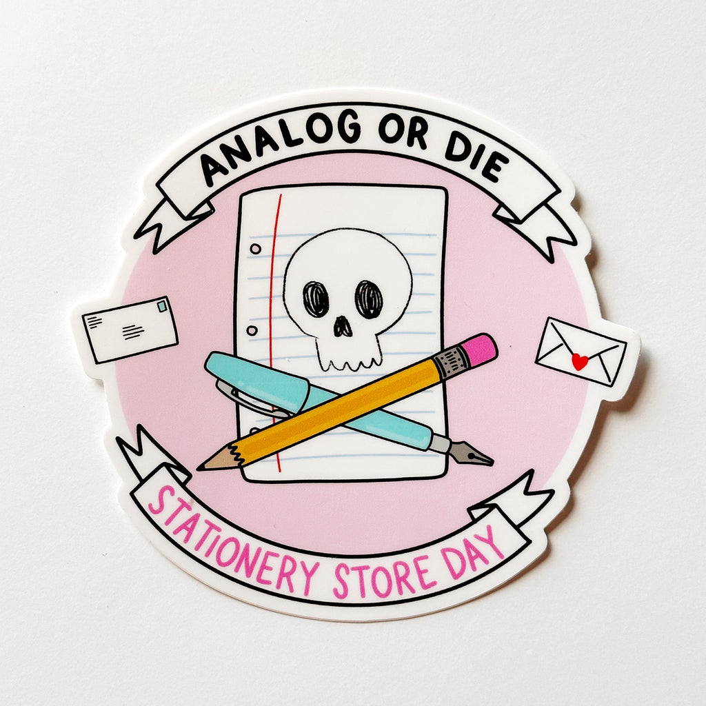 Image of pink circle sticker with image of white skull on a piece of notebook paper with blue pen and yellow pencil crossed  and black text at top says, "Analog or die" and pink text says, "Stationery Store Day". 