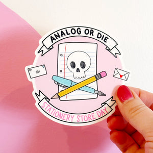 Image of pink circle sticker with image of white skull on a piece of notebook paper with blue pen and yellow pencil crossed and black text at top says, "Analog or die" and pink text says, "Stationery Store Day".
