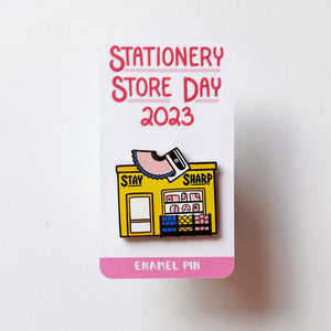 Image of enamel pin of yellow storefront with pencil sharpener and black text says, "Stay sharp". 