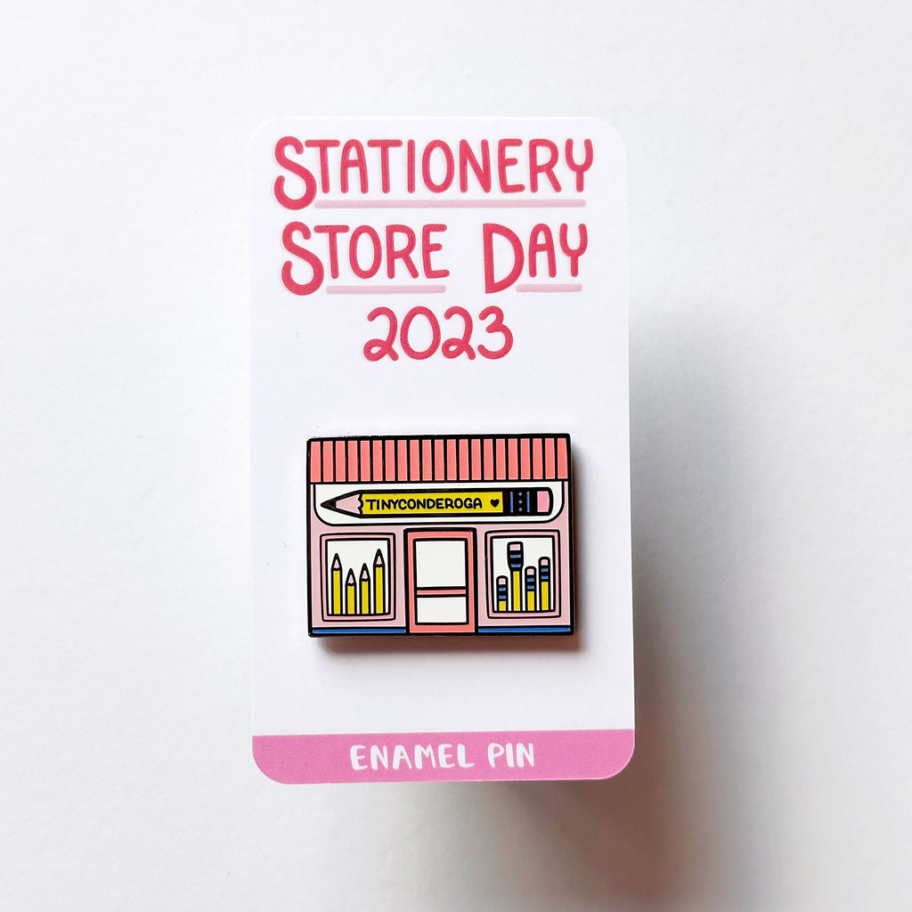 Image of enamel pin of pink storefront with pencil sign says "Tinyconderoga" in black text. 