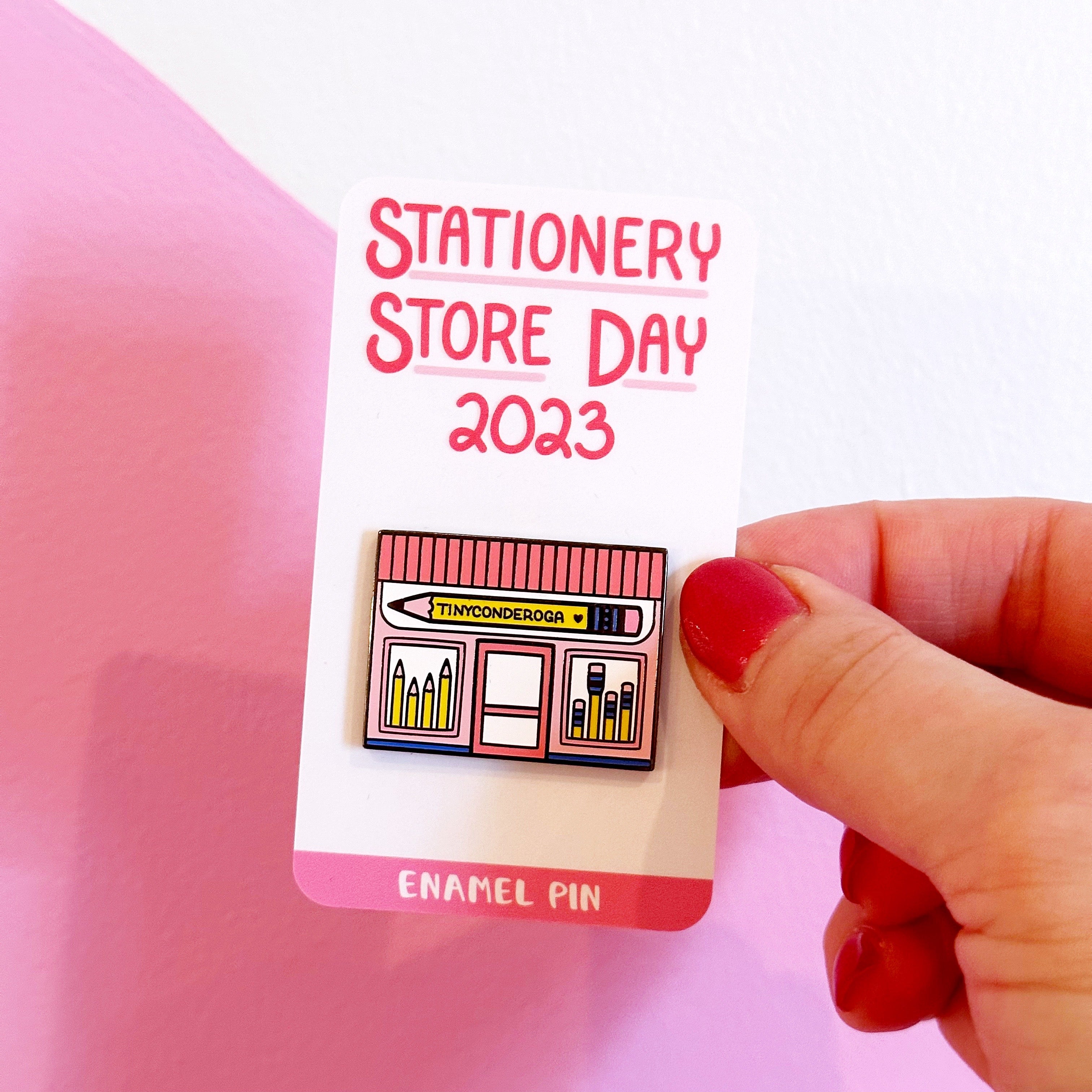 Image of enamel pin of pink storefront with pencil sign says "Tinyconderoga" in black text.