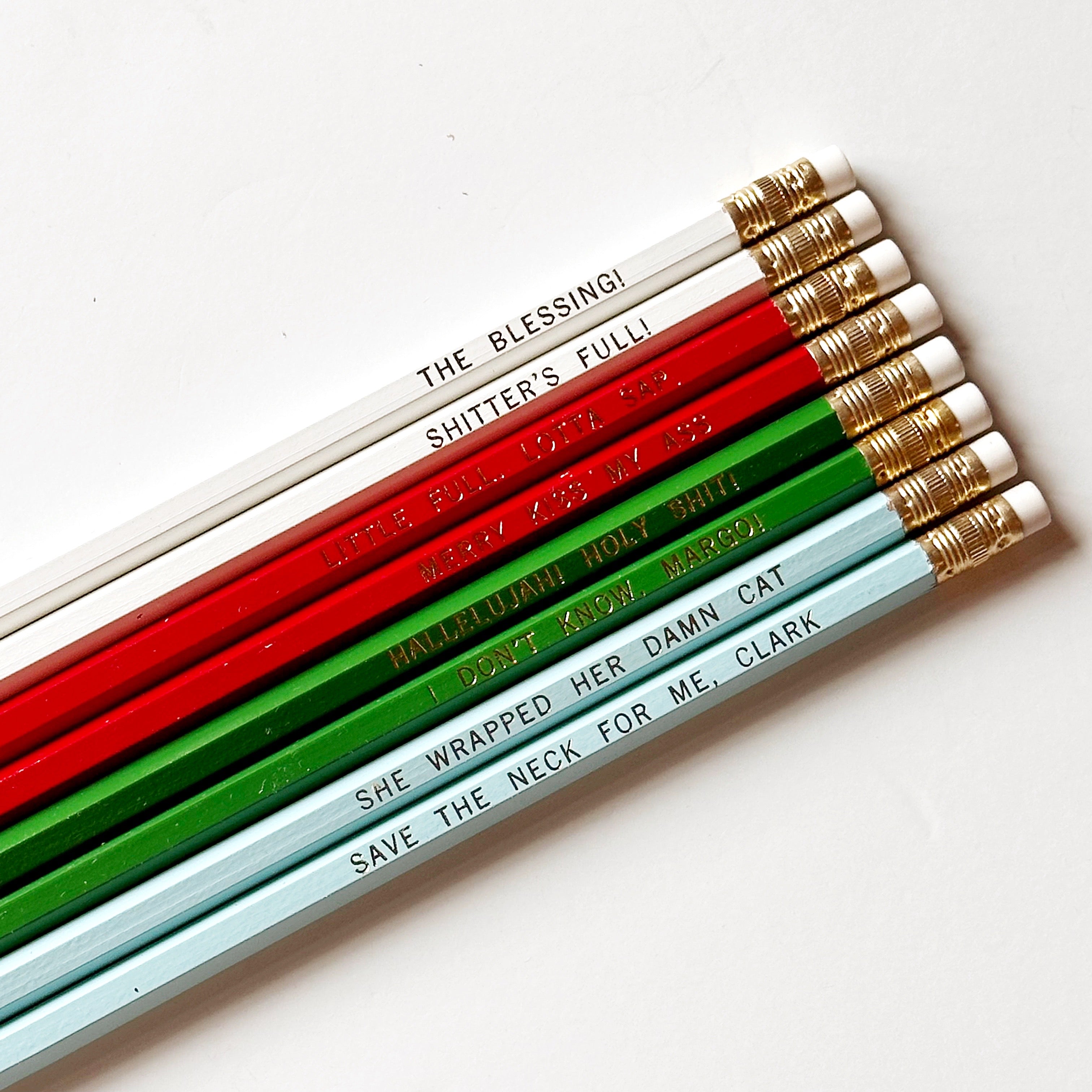 Eight pencils with gold foil text says, "Merry Kiss My Ass Little Full. Lotta Sap. The Blessing! Shitter's Full! Hallelujah! Holy Shit! I Don't Know, Margo! She Wrapped Her Damn Cat Save The Neck For Me, Clark". 